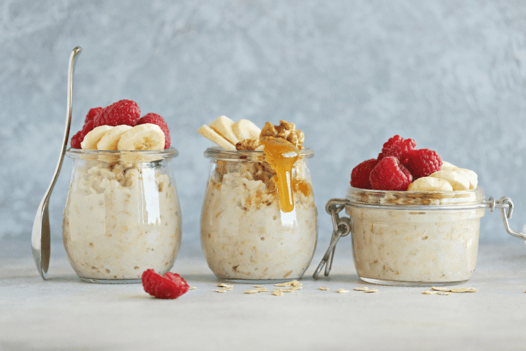 Three glass jars of mixed berry cheesecake overnight oats topped with banana, raspberries, and honey – a healthy no-bake breakfast idea.